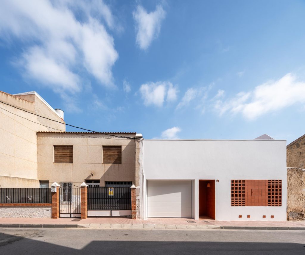 `Fachada exterior de Casa tras celosías, vivienda entre medianeras diseñada por Nítida arquitectos en Murcia, con volumen blanco, celosía de barro y acceso en naranja`